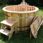 A wooden hot tub with metal bands sits on a grassy lawn, with a small wooden step stool nearby.