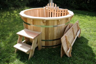 A wooden hot tub with metal bands sits on a grassy lawn, with a small wooden step stool nearby.