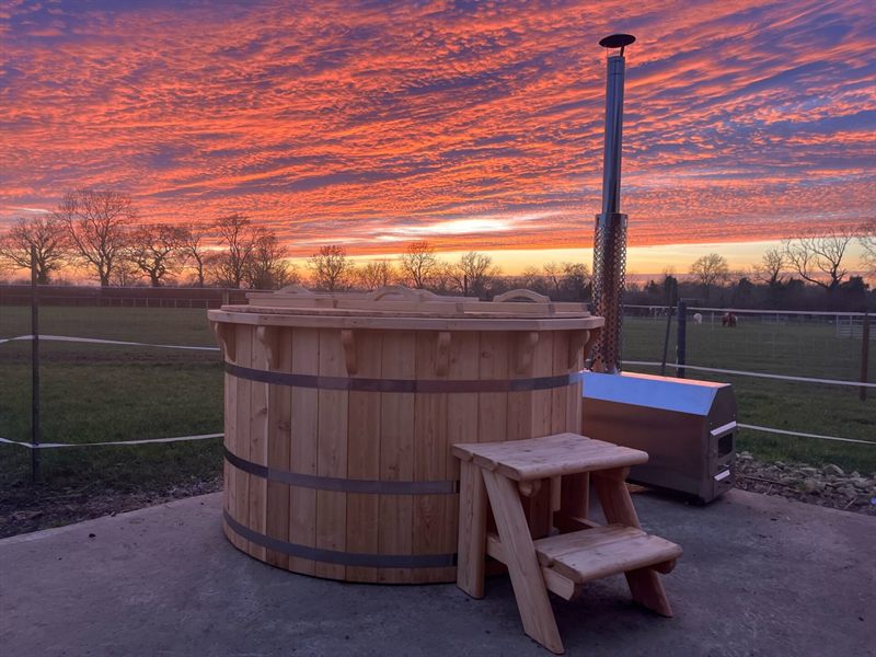 Wooden hot tub with a wooden step stool beside it, metal chimney, and sunset sky.