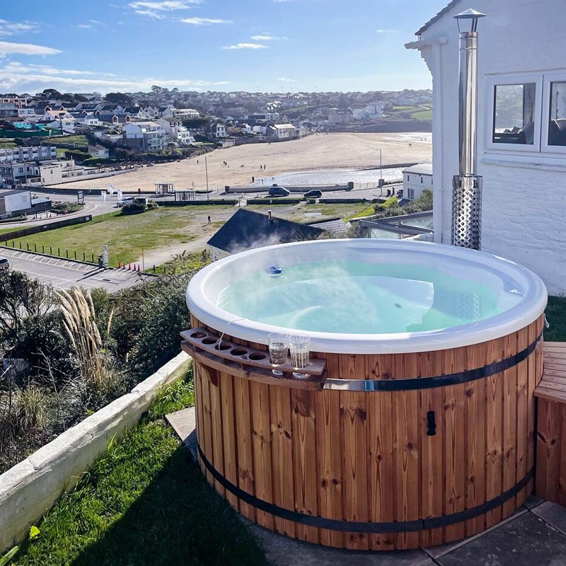 Round wooden hot tub on a patio overlooking a coastal town and beach.