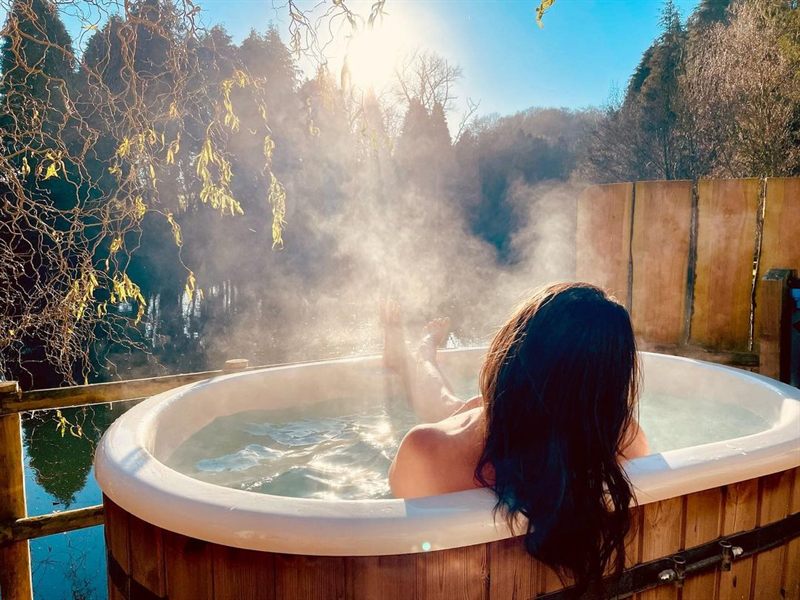 Person relaxing in an outdoor wooden hot tub, steam rising, back to camera, legs up, surrounding trees and fence in bright sunlight.